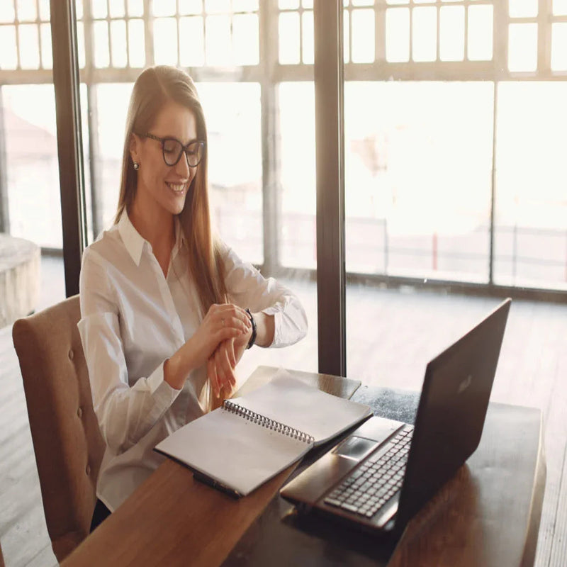 Smiling woman in glasses checking smartwatch at desk with laptop and open notebook in bright office