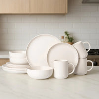 Full 12-piece off-white matte stoneware dinnerware set arranged on white marble kitchen counter with wood cabinetry background