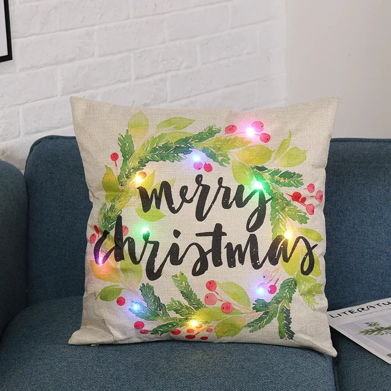 Decorative beige Christmas pillow with green wreath, red berries, and colorful LED lights on blue sofa