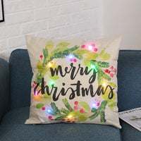 Decorative beige Christmas pillow with green wreath, red berries, and colorful LED lights on blue sofa