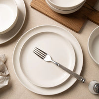 Top-down view of off-white matte stoneware dinner plate and salad plate stacked with fork, acacia wood board and cereal bowls in background