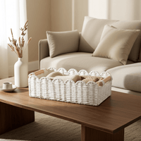 White paper rope woven storage basket with wood handles filled with a folded knit throw blanket, placed on a wood coffee table beside a beige linen sofa with pillows, dried flowers in a white ceramic vase and a small ceramic dish nearby, warm natural sunlight.