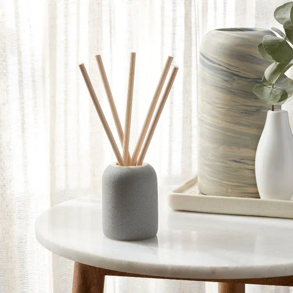 Gray ceramic reed diffuser placed on marble side table with white curtains and decorative vase in cozy living room