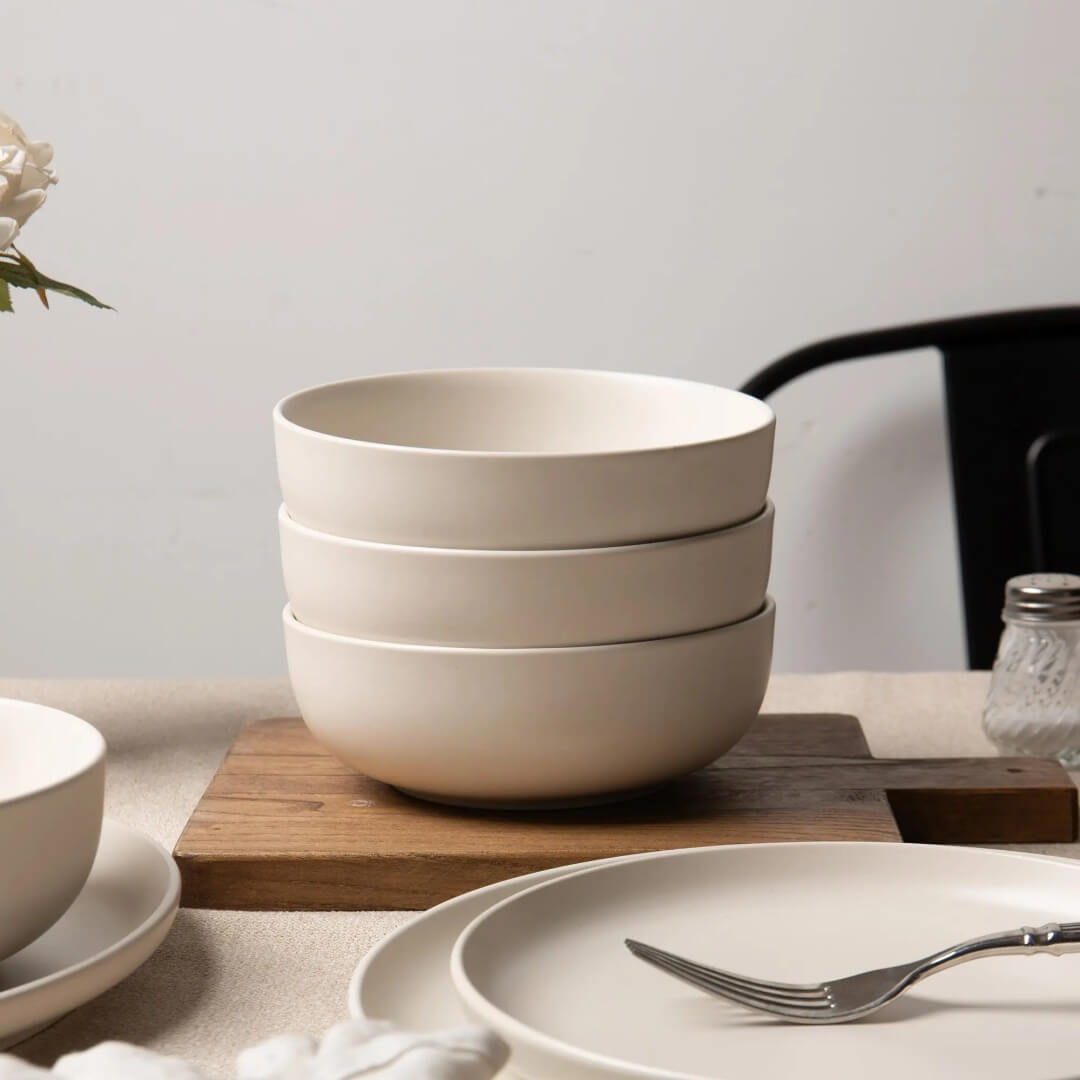 Three off-white matte stoneware cereal bowls stacked on acacia wood board with dinner plate and fork in foreground