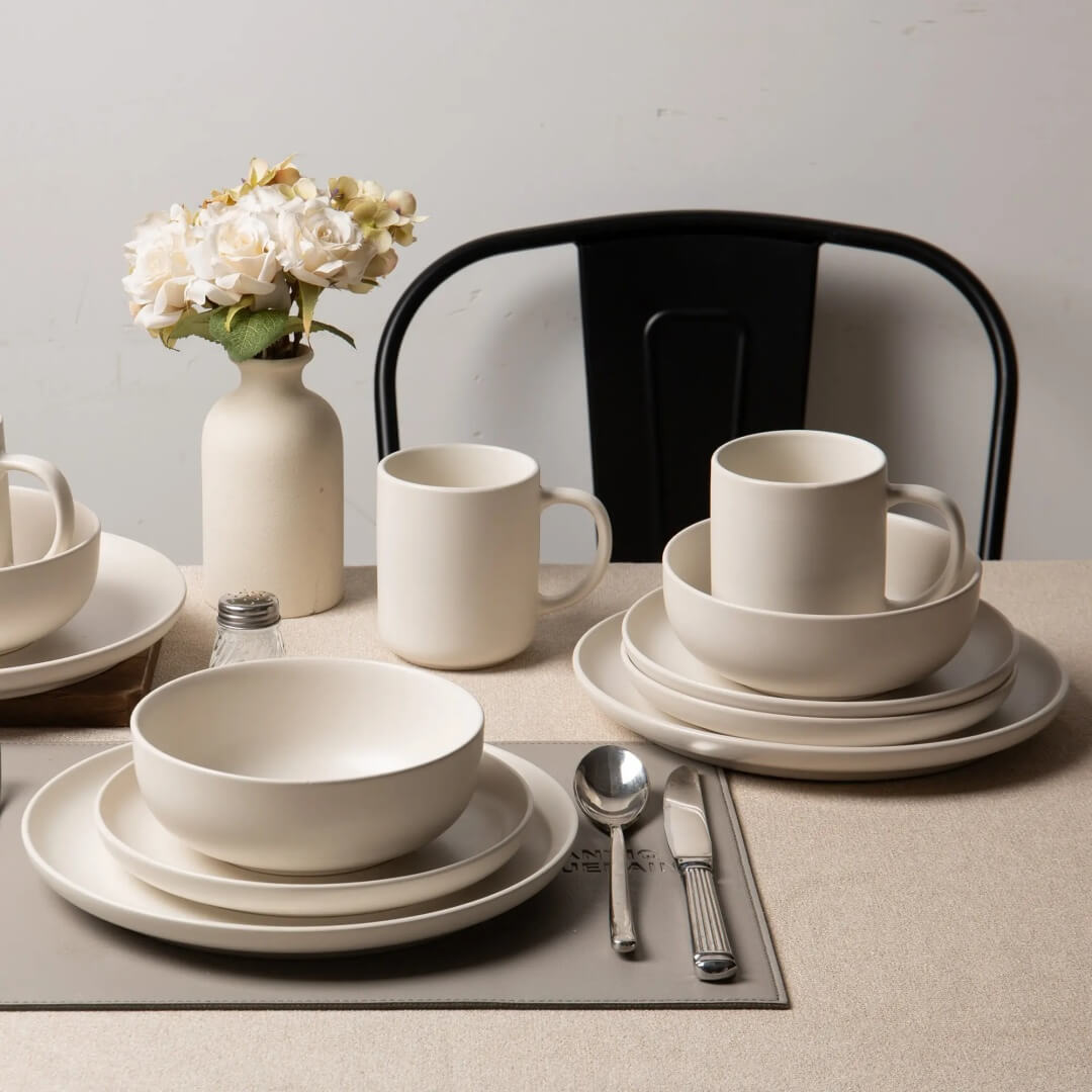Two place settings with matte stoneware bowls, plates and mugs on linen tablecloth beside white ceramic vase with flowers and black metal chair