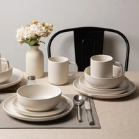 Two place settings with matte stoneware bowls, plates and mugs on linen tablecloth beside white ceramic vase with flowers and black metal chair