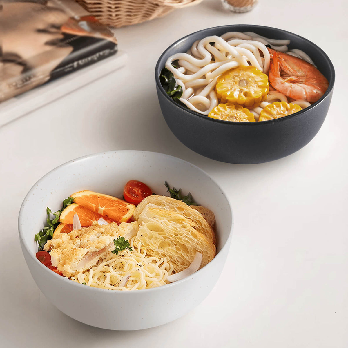 White speckled and dark charcoal matte ceramic bowls side by side filled with noodle dishes on white table
