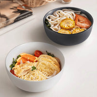 White speckled and dark charcoal matte ceramic bowls side by side filled with noodle dishes on white table