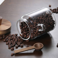 Glass coffee jar tilted showing coffee beans with wooden spoon and glass handle