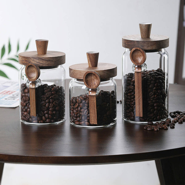 Three glass coffee jars with walnut wood lids and spoons filled with coffee beans on table