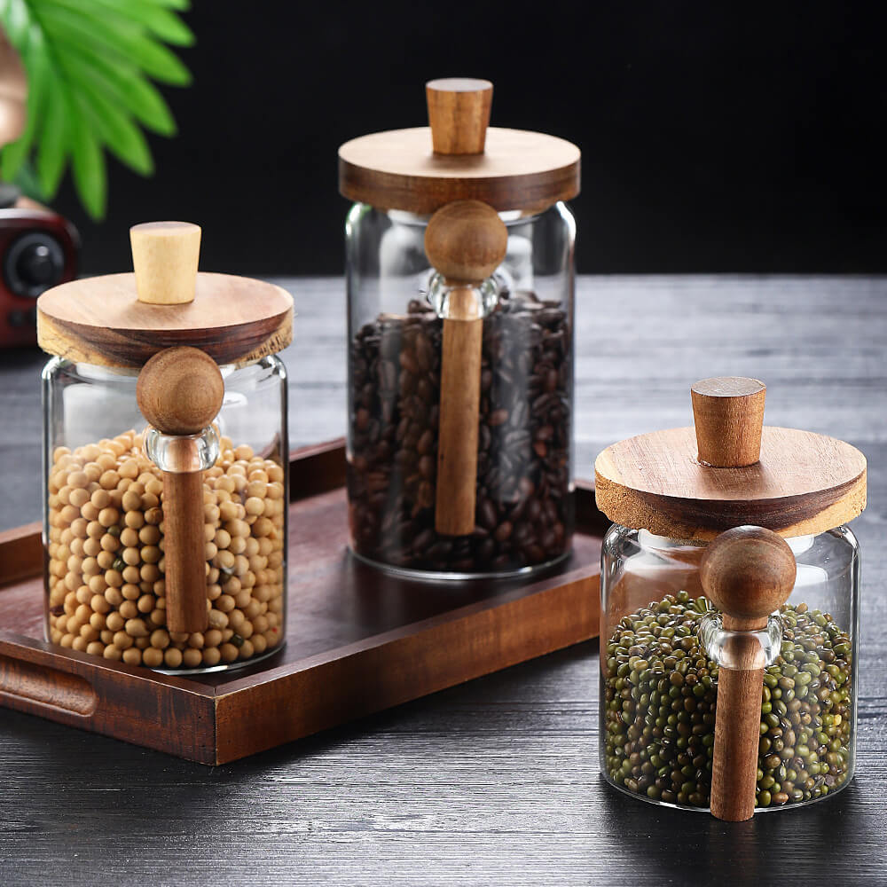 Set of 3 borosilicate glass storage jars with natural wooden lids and spoons on dark surface